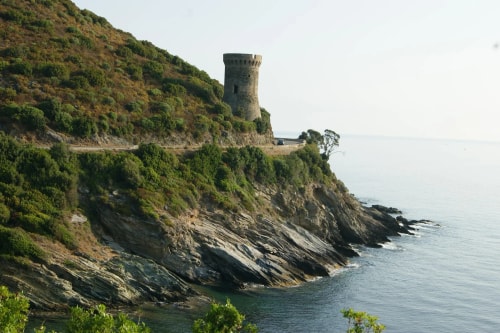 Une tour en pierre perchée sur une falaise, surplombant la mer, entourée de verdure et de rochers.