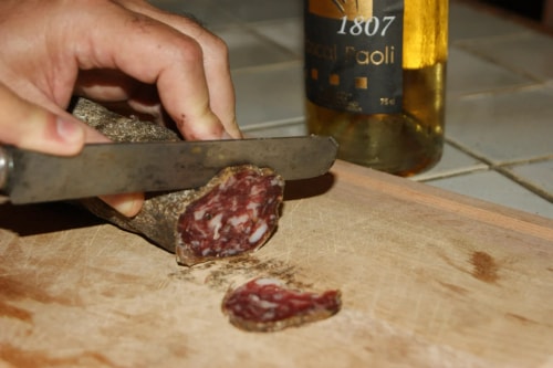 Une main coupant un saucisson sur une planche à découper, avec une bouteille de vin en arrière-plan.