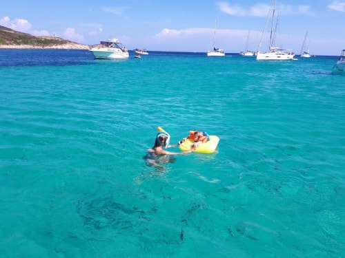 Une scène de plage avec une personne nageant dans une eau turquoise, équipée d'un masque de plongée, s'amusant avec un enfant dans une bouée flottante. En arrière-plan, on peut voir des bateaux à moteur sur l'eau.