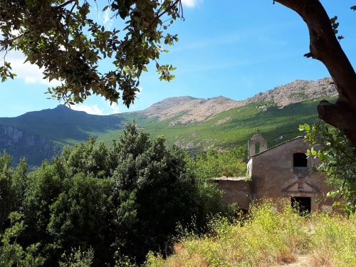 Vue d'une maison en ruine entourée d'arbres et de montagnes verdoyantes sous un ciel bleu.
