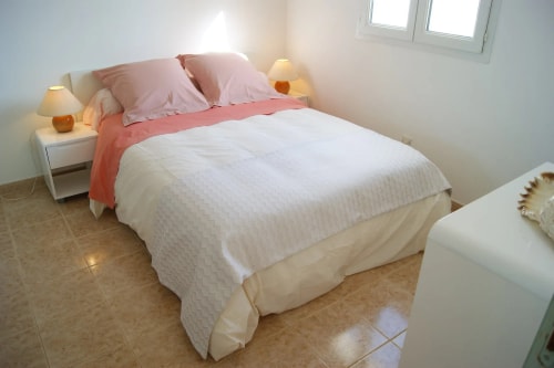 Chambre à coucher avec un lit double, draps blancs et oreillers roses, lampes de chevet sur des tables blanches, sol carrelé