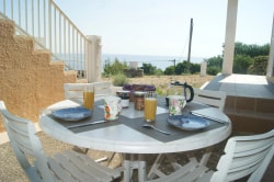 Table dressée avec des assiettes, des verres de boisson et des couverts, devant une vue sur la mer, avec des chaises blanches, dans un environnement extérieur ensoleillé.