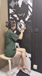 Une femme dessine sur un mur noir décoré d’un visage abstrait en blanc, dans une pièce aux murs lambrissés et au sol carrelé.
