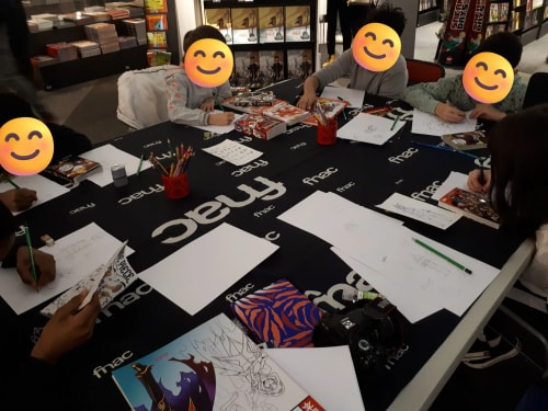 Un groupe d'enfants participe à un atelier de dessin autour d'une grande table noire marquée du logo Fnac, avec des feuilles, des crayons et des mangas. Leurs visages sont masqués par des émojis souriants.