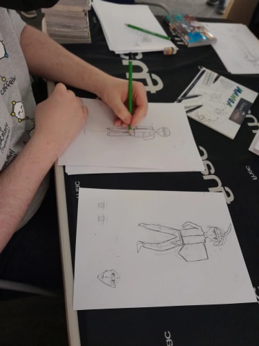 Un enfant dessine des personnages de style manga au crayon sur des feuilles de papier posées sur une table sombre, avec des exemplaires de livres et du matériel de dessin autour.