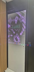 Écran vertical affichant une interface musicale violette « Lutti », avec formes d’ondes, illustration stylisée et commandes de lecture, dans un intérieur aux panneaux en bois.