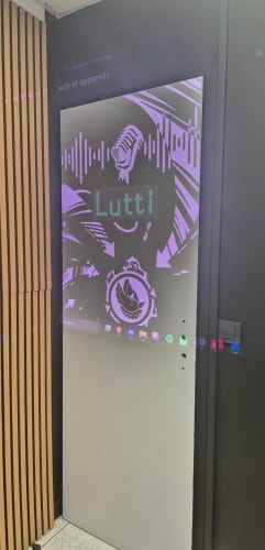 Écran vertical affichant une interface musicale violette « Lutti », avec formes d’ondes, illustration stylisée et commandes de lecture, dans un intérieur aux panneaux en bois.