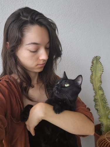 Une femme aux cheveux longs tient un chat noir dans ses bras, près d’un cactus en pot, sur fond de mur gris.