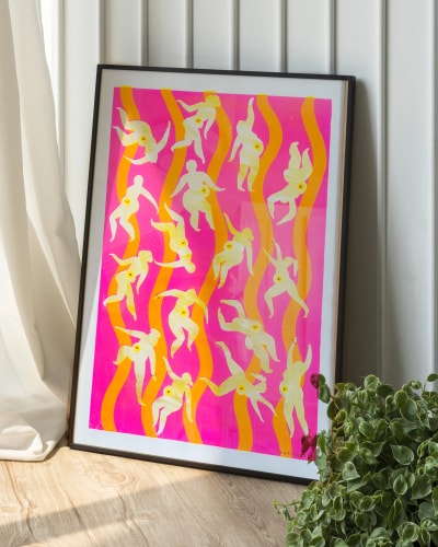Affiche encadrée posée au sol, montrant des silhouettes féminines stylisées jaune pâle sur un fond rose vif orné de lignes verticales ondulées orange, avec une plante verte à côté.