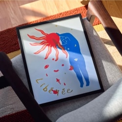 Affiche encadrée posée sur une chaise, montrant une silhouette bleue touchant un soleil rouge avec le mot « Libérée » écrit en lettres dorées, entouré de petits symboles mystiques sur fond blanc.