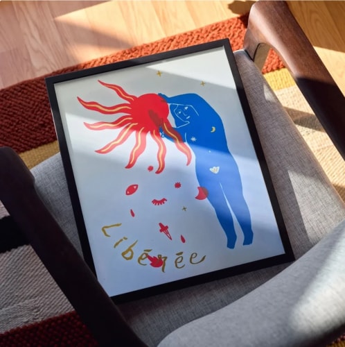 Affiche encadrée posée sur une chaise, montrant une silhouette bleue touchant un soleil rouge avec le mot « Libérée » écrit en lettres dorées, entouré de petits symboles mystiques sur fond blanc.