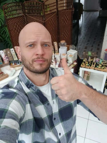 Charles-Henri. Homme chauve portant une chemise à carreaux blancs et verts faisant un pouce levé devant une table avec divers produits exposés.
