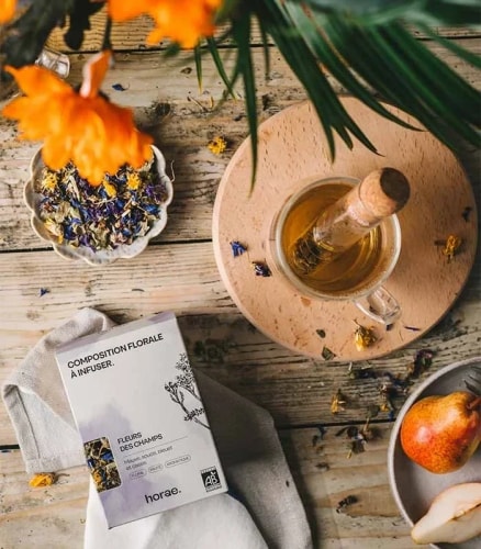 Composition florale à infuser avec infusion dans une tasse en verre sur un support en bois, fleurs séchées dans un petit plat, une poire entière et une tranche de poire sur une assiette, le tout posé sur une table en bois rustique.