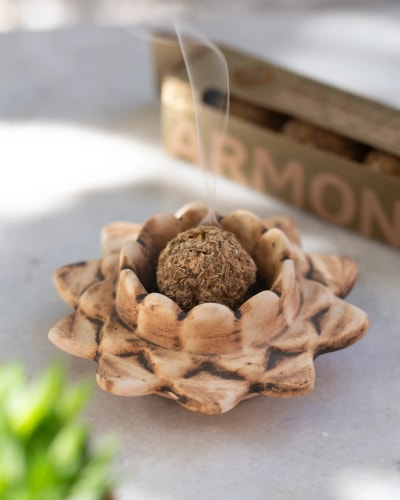 Boule d'encens allumée fumante placée au centre d'un porte-encens en forme de fleur en céramique marron clair, sur une surface claire avec un paquet d'encens en arrière-plan.