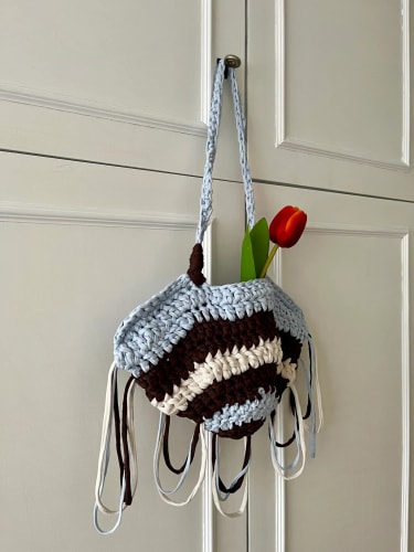 Un sac au crochet rayé bleu, blanc et marron avec des franges suspendu à une poignée de porte, contenant une tulipe rouge.