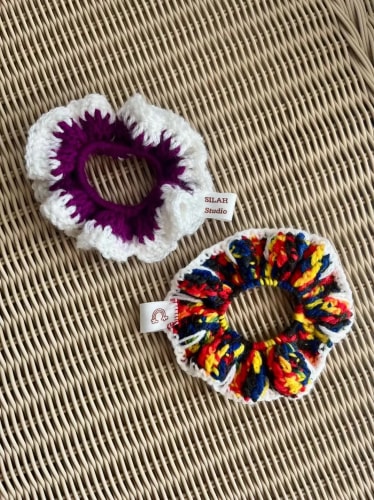 Deux chouchous en crochet faits main posés sur une surface en osier. L'un est violet et blanc, l'autre est multicolore avec des touches de rouge, bleu et jaune, chacun avec une petite étiquette textile.