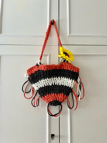 Sac en crochet rayé orange, noir et blanc, agrémenté de franges et d'un tournesol décoratif, suspendu devant une porte à panneaux blancs.