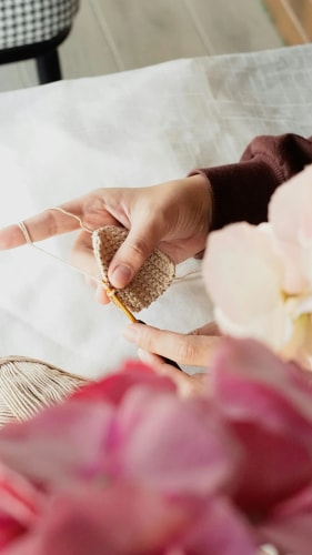 Gros plan sur des mains en train de crocheter un ouvrage en fil beige. Au premier plan, des fleurs roses sont légèrement floues. En arrière-plan, une surface claire et un bout de chaise à carreaux.