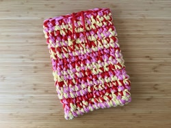 Une pochette d'ordinateur rectangulaire crochetée en bandes de tissu rose, jaune et rouge, posé sur un sol en bois clair.