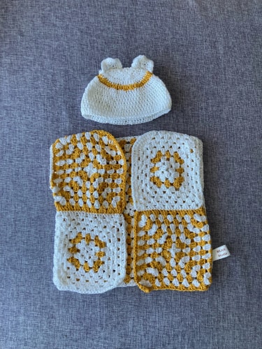 Ensemble pour bébé au crochet blanc et jaune moutarde composé d'un bonnet à petites oreilles et d'un petit haut fait de carrés granny sur un fond gris.