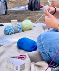 Pelote de laine bleue au premier plan, pelote verte et fils colorés sur une couverture, avec des mains tenant des aiguilles à crochet.