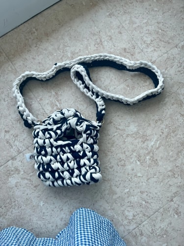 Petit sac à main crocheté avec un motif bicolore en bleu nuit et blanc, doté d'une longue bandoulière assortie, posé sur un sol carrelé beige.