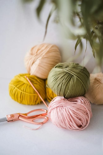 Pelotes de laine de couleurs douces (jaune, rose, vert, beige) posées sur une table à côté d'une paire de ciseaux en métal cuivré, avec des feuilles d'eucalyptus en arrière-plan.