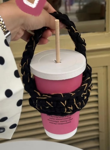 Une main tient un gobelet rose avec une paille, inséré dans un porte-gobelet en crochet noir tressé.