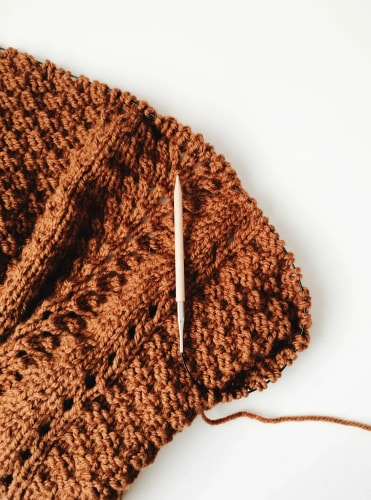 Un tricot en laine de couleur marron avec une aiguille en bois posée dessus, sur fond blanc.