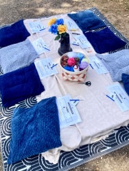 Atelier crochet au sol avec nappe blanche, coussins bleus, fleurs, panier de fruits rouges et cartes disposés sur un tapis à motifs.