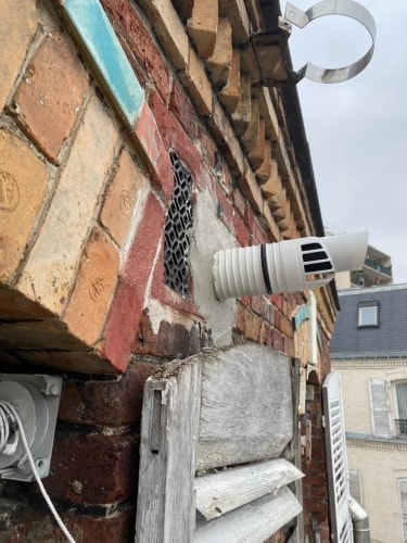 Mur en briques anciennes avec une sortie de conduit de cheminée blanche et une grille de ventilation, fixé sur une façade au-dessus d'un volet en bois.