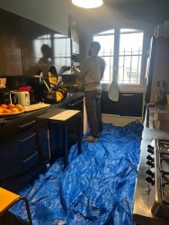 Un homme debout dans une cuisine travaille sur une installation murale, le sol est recouvert d'une bâche de protection bleue.