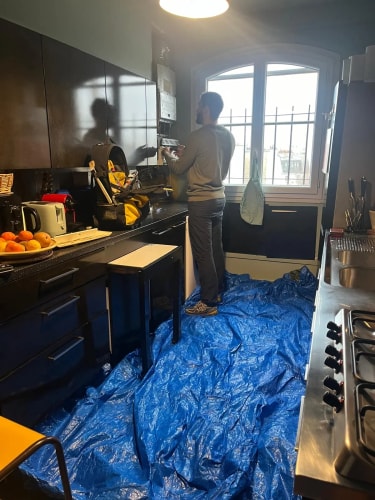 Un homme debout dans une cuisine travaille sur une installation murale, le sol est recouvert d'une bâche de protection bleue.