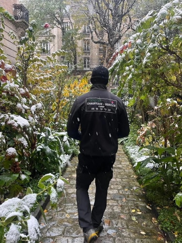 Un homme vêtu de noir marche sur un chemin pavé dans un jardin sous la neige, entouré de plantes et d'arbres partiellement enneigés avec un immeuble en arrière-plan.