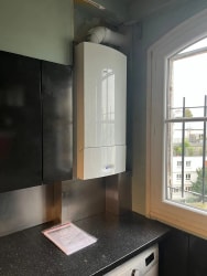 Chaudière murale blanche installée dans une cuisine, au-dessus d'un plan de travail, à côté d'une fenêtre et de placards noirs.