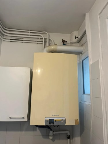 Chaudière murale beige installée sur un mur blanc, avec des tuyaux de raccordement visibles au-dessus et un petit placard blanc à gauche.