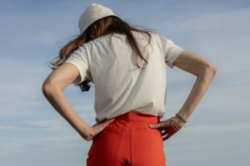 Une femme de dos portant le bob cloche écru et le short rouge coquelicot de la marque avec un ciel marin en fond