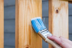 Une main tenant un pinceau avec des poils bleus peignant une surface en bois clair, pour illustrer le fait de redonner un coup de neuf