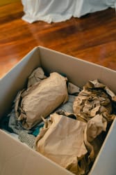 Une boîte en carton remplie de papier froissé et de vêtements pliés, sur un sol en bois, pour illustrer le fait de désencombrer son intérieur