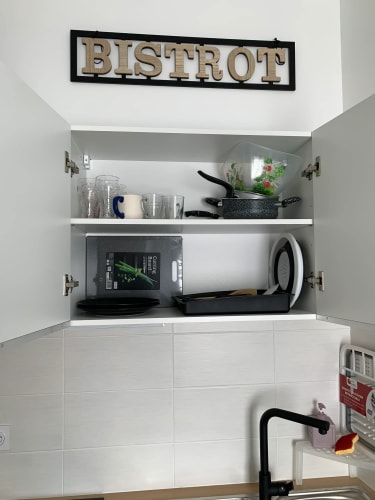 Placard de cuisine ouvert avec vaisselle et ustensiles, notamment verres, tasses, casseroles, saladier, planche à découper et plateaux, avec un panneau décoratif au-dessus portant l'inscription «BISTROT» par Inès Home Design