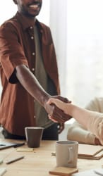 Deux personnes se serrent la main, l'une avec une chemise marron et l'autre portant un pull clair, sur une table avec des tasses, pour illustrer la validation des idées avec la cliente ou le client