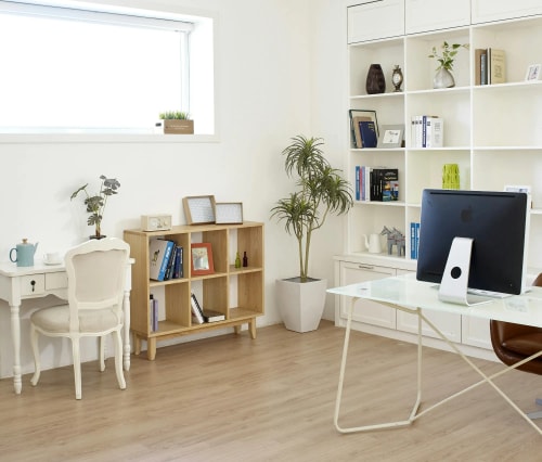 Intérieur de bureau moderne avec une table de travail en verre, un ordinateur sur un support, des étagères blanches remplies de livres, une plante en pot et un bureau avec une chaise élégante.