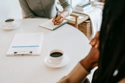 Une table ronde avec deux tasses de café, une personne prenant des notes dans un carnet et une autre personne, floue, les mains posées sur la table, pour illustrer la première étape d'échange