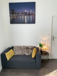 Petit canapé gris avec coussins jaunes et couverture en fausse fourrure, table d'appoint noire avec lampe allumée et plante verte, tableau représentant un pont et une ville la nuit accroché au mur blanc par Inès Home Design
