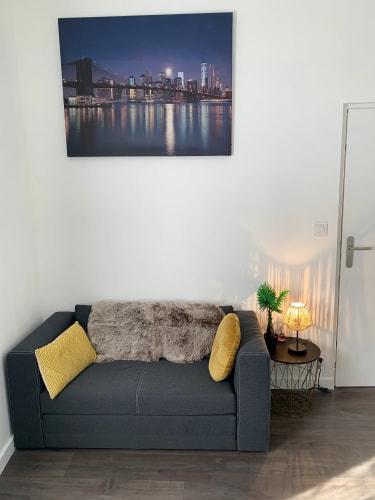 Petit canapé gris avec coussins jaunes et couverture en fausse fourrure, table d'appoint noire avec lampe allumée et plante verte, tableau représentant un pont et une ville la nuit accroché au mur blanc par Inès Home Design