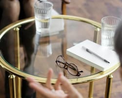 Une table en verre avec des lunettes posées dessus, un carnet blanc et un stylo, entourée de verres d'eau, pour illustrer le service de relecture et réécriture par Les Belles Personnes