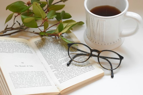 Une tasse de café posée à côté d'un livre ouvert, avec des lunettes noires et une branche de feuilles vertes, pour illustrer le service de lecture de manuscrit proposé par l'agence éditoriale Les Belles Personnes