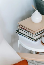 Une table blanche avec une pile de livres et un vase blanc contenant une tige, sur un fond clair.