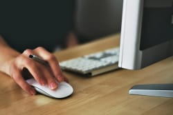 Main gauche tenant une souris d'ordinateur avec un stylo dans l'autre main, devant un clavier et un écran sur un bureau en bois, pour illustrer le service de présentation de manuscrit à des maisons d'édition par Les Belles Personnes
