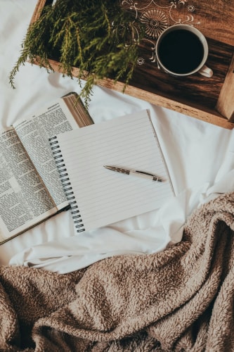 Une vue du dessus d'un lit avec un drap blanc, un livre ouvert, un carnet à spirale blanc avec un stylo posé dessus, et une tasse de café sur un plateau en bois, pour illustrer le service le lecture et annotation des Belles Personnes